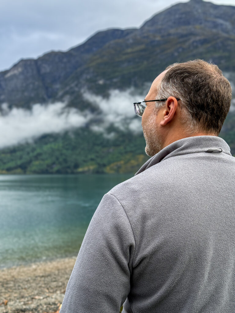 Man standing along the shoreline of a Norwegian fjord with mountains and mist-covered slopes in the background.