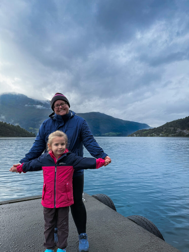 Mother and daughter standing on a dock overlooking a scenic Norwegian fjord surrounded by mountains and low clouds.